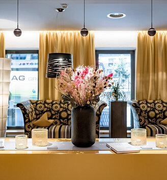 A modern reception room with yellow curtains, patterned sofas, pendant lights and a table holding a black vase with pink flowers, candles and magazines. Large windows let in daylight.