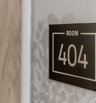 A sign reading "Room 404" in white lettering on a black background, hangs on the wall next to a light-colored wooden door and patterned wallpaper.