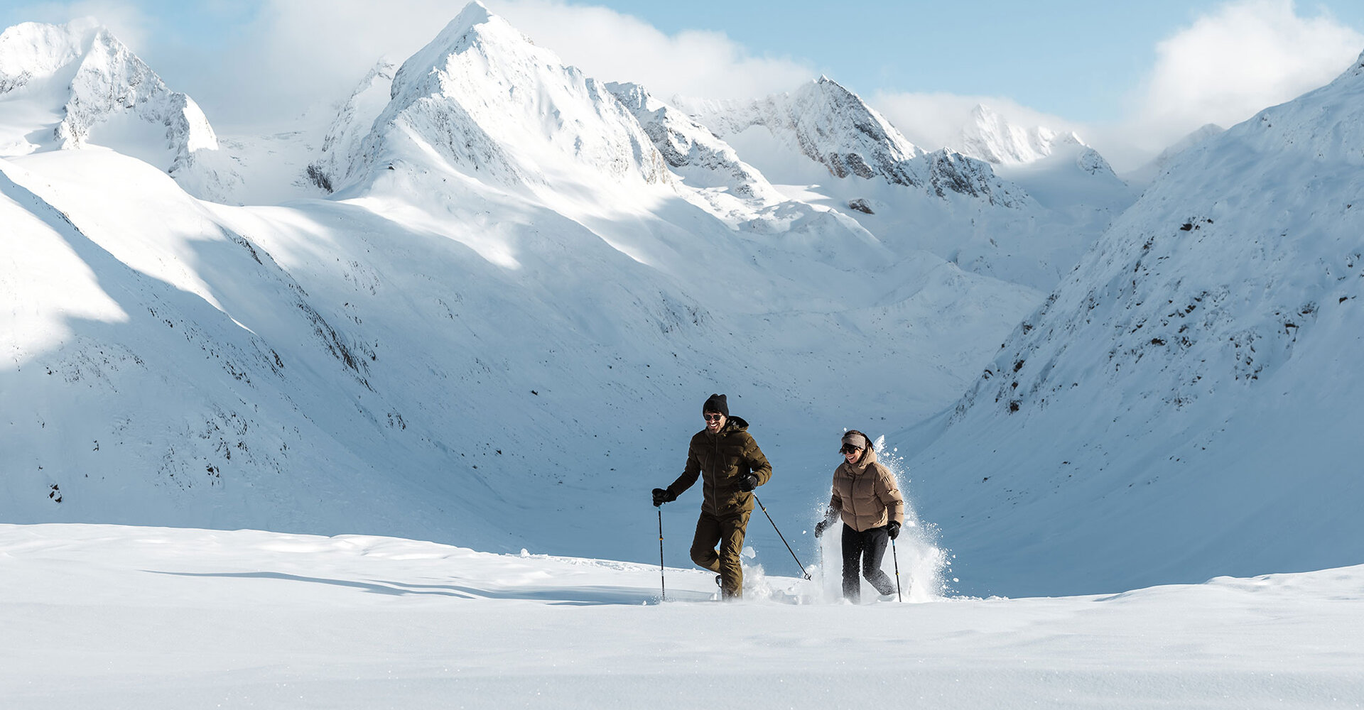 Two people are hiking on snowshoes through the deep snow in a snow-white mountain landscape with high, snow-covered peaks under a clear sky.