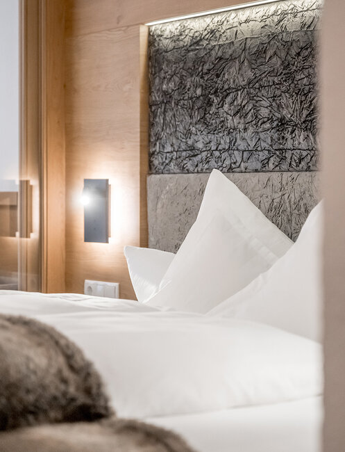 A modern hotel room featuring a neatly made bed, white pillows, a fluffy blanket, wood accents and a textured headboard. Soft lighting creates a cozy atmosphere.