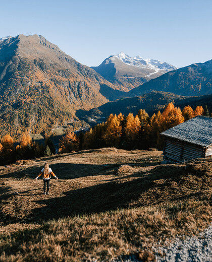 A person walks across a grassy slope dotted with wooden huts, surrounded by trees in autumn colors and snow-capped mountains under a brilliant blue sky.