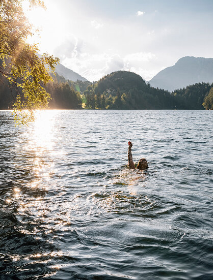 Eine Person, die in einem See schwimmt und einen Arm über das Wasser hebt, während sich das Sonnenlicht auf dem Wasser spiegelt und im Hintergrund bewaldete Berge zu sehen sind.