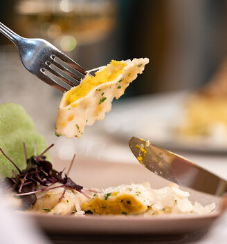 A person is eating ravioli from a plate with a fork and knife: the dish is served with microgreens and a green side dish. A glass of white wine is in the background.