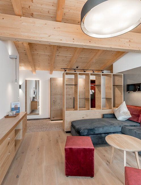 Modernes Wohnzimmer mit Holzbalken an der Decke, einem an der Wand montierten Fernseher, einem Ecksofa mit roten und grauen Kissen, einem kleinen runden Tisch und einem Holzschrank. Im Hintergrund sind ein Spiegel und offene Regale zu sehen.
