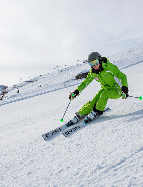 A skier in a bright green outfit and a helmet speeds down a snowy mountain slope, leaning into a turn with ski poles in hand. The sky is partly cloudy with mountains in the distance.