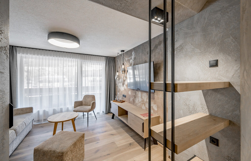Modern living room with light wood floors, a beige sofa and an armchair. A round coffee table, a wall-mounted TV and a large window with sheer curtains complete the minimalist décor. Shelving units are located on the right side.