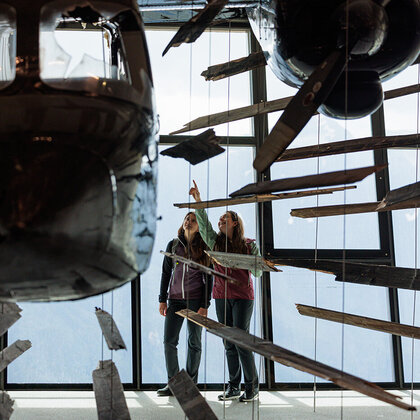 Two people are standing in a museum, looking at a suspended, fragmented installation in the shape of an airplane while pointing at it. The entire scene is set against large windows.