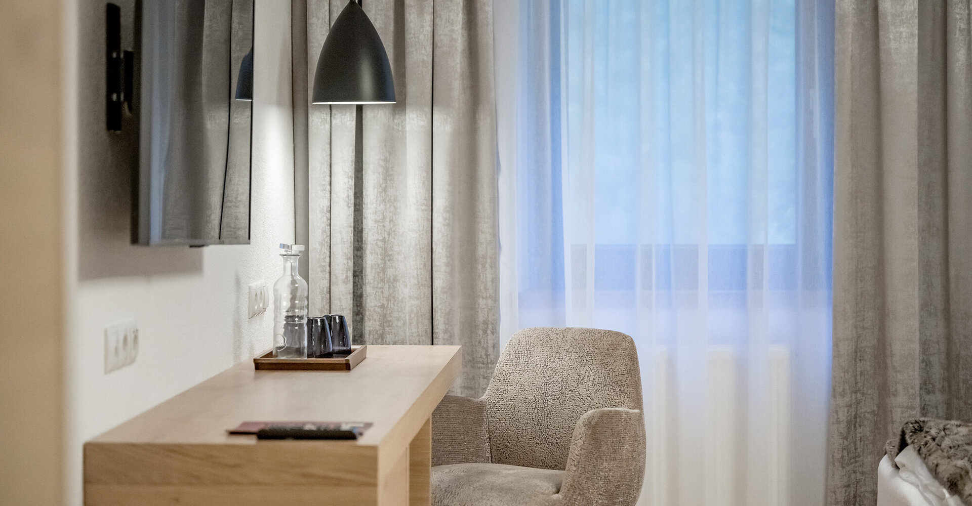 A modern hotel room with a light-wood desk, an upholstered chair, a wall-mounted TV, a pendant light and sheer curtains over the window.