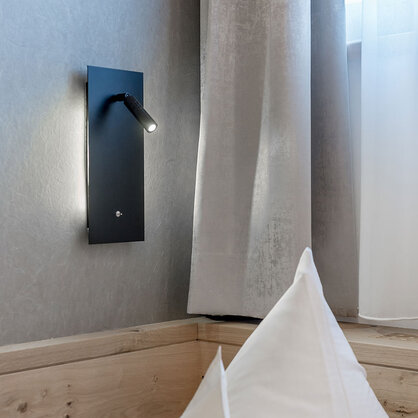Next to a bed with white pillows, near a window with sheer and gray curtains, a modern black wall-mounted reading lamp is switched on.