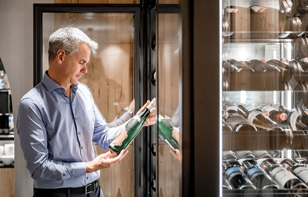 A grey-haired man in a light blue shirt examines a wine bottle in a modern setting, standing in front of a glass wine cooler filled with bottles.