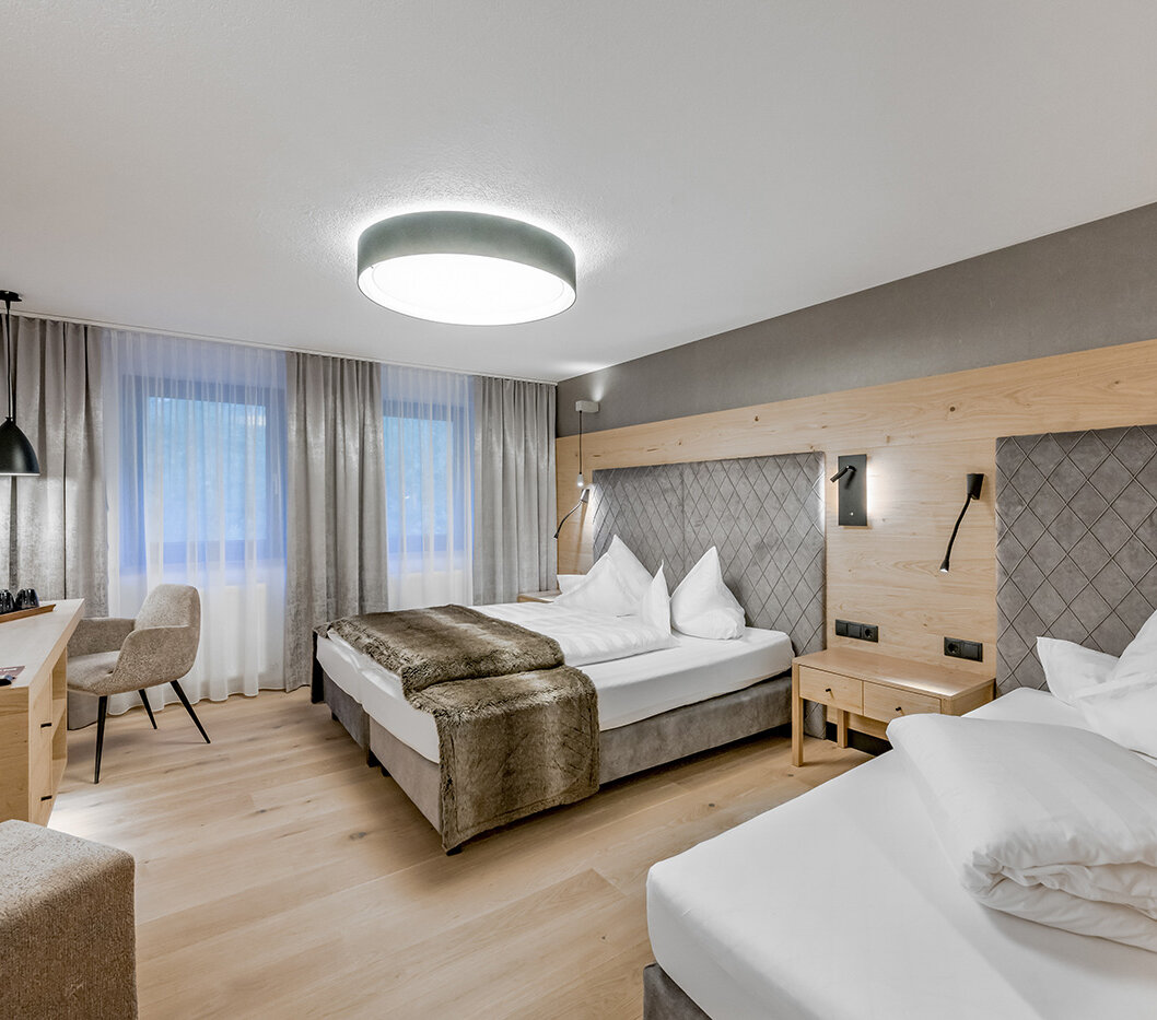 A modern hotel room featuring two beds, a desk with a chair, a wall-mounted TV, a large window with curtains and soft lighting. The décor highlights wood accents and neutral tones, creating a cozy and inviting atmosphere.
