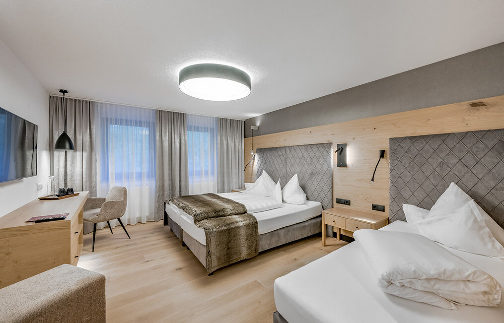 A modern hotel room featuring two beds, a desk with a chair, a wall-mounted TV, a large window with curtains and soft lighting. The décor highlights wood accents and neutral tones, creating a cozy and inviting atmosphere.