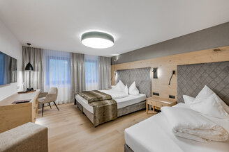 A modern hotel room featuring two beds, a desk with a chair, a wall-mounted TV, a large window with curtains and soft lighting. The décor highlights wood accents and neutral tones, creating a cozy and inviting atmosphere.