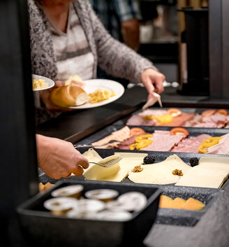 People helping themselves to cheese, cold cuts and other foods at the buffet. With plates in hand and under bright lighting, they choose from various types of meat, cheese slices and garnished dishes.