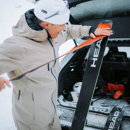 A person dressed in winter clothing attaches a ski touring skin to a pair of skis in a snowy landscape, next to the open trunk of a car.