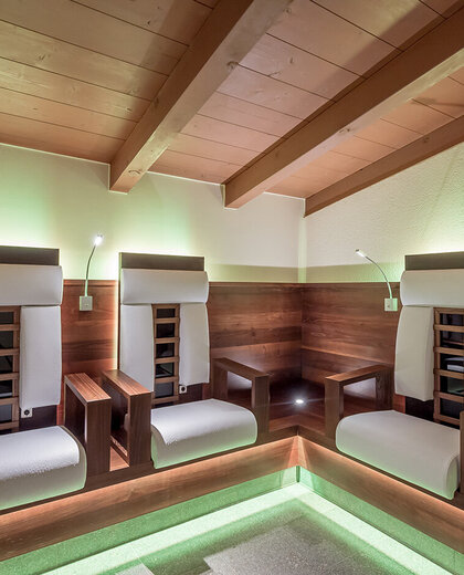 Three modern infrared armchairs with white cushions are situated in a cozy corner room featuring exposed wooden beams, soft green accent lighting and reading lamps above each seat.