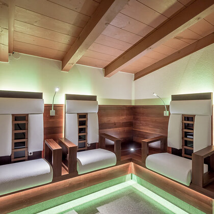 Three modern infrared armchairs with white cushions are situated in a cozy corner room featuring exposed wooden beams, soft green accent lighting and reading lamps above each seat.