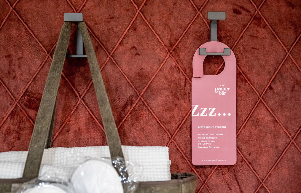 A pink door hanger and a bag containing towels hang on hooks in front of a red quilted wall.