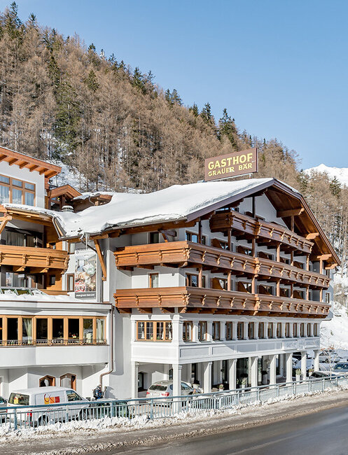 A large hotel with wooden balconies stands on a snow-covered road, surrounded by snow-capped trees and mountains under a brilliant blue sky.