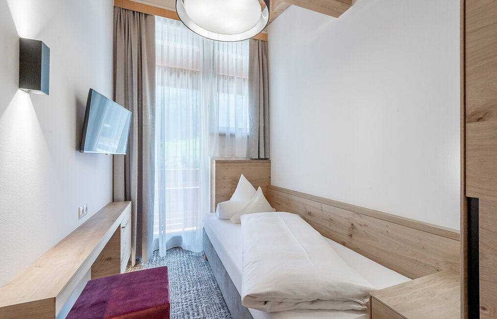 A small, modern room with a single bed, a wall-mounted TV, a shelf and a floor-to-ceiling window with sheer curtains that let daylight stream in.