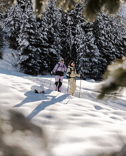 Two people in winter clothing stand amidst a snowy landscape, surrounded by snow-covered trees. Sunlight shines through the fir branches in the foreground, creating a picturesque winter atmosphere.