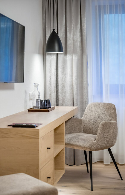 Ein gemütlicher Arbeitsbereich mit einem beigen Stuhl, einem Schreibtisch aus Holz, einem Tablett mit Gläsern und einer schwarzen Pendelleuchte. Im Hintergrund sind graue Vorhänge und ein Fenster mit weißen Gardinen zu sehen.
