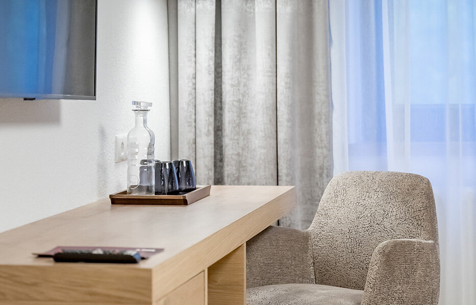 A cozy workspace featuring a beige chair, a wooden desk, a tray with glasses and a black pendant light. In the background, there are gray curtains and a window with white sheer curtains.