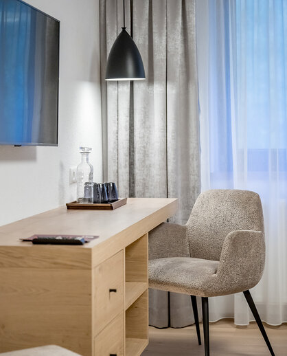 A cozy workspace featuring a beige chair, a wooden desk, a tray with glasses and a black pendant light. In the background, there are gray curtains and a window with white sheer curtains.