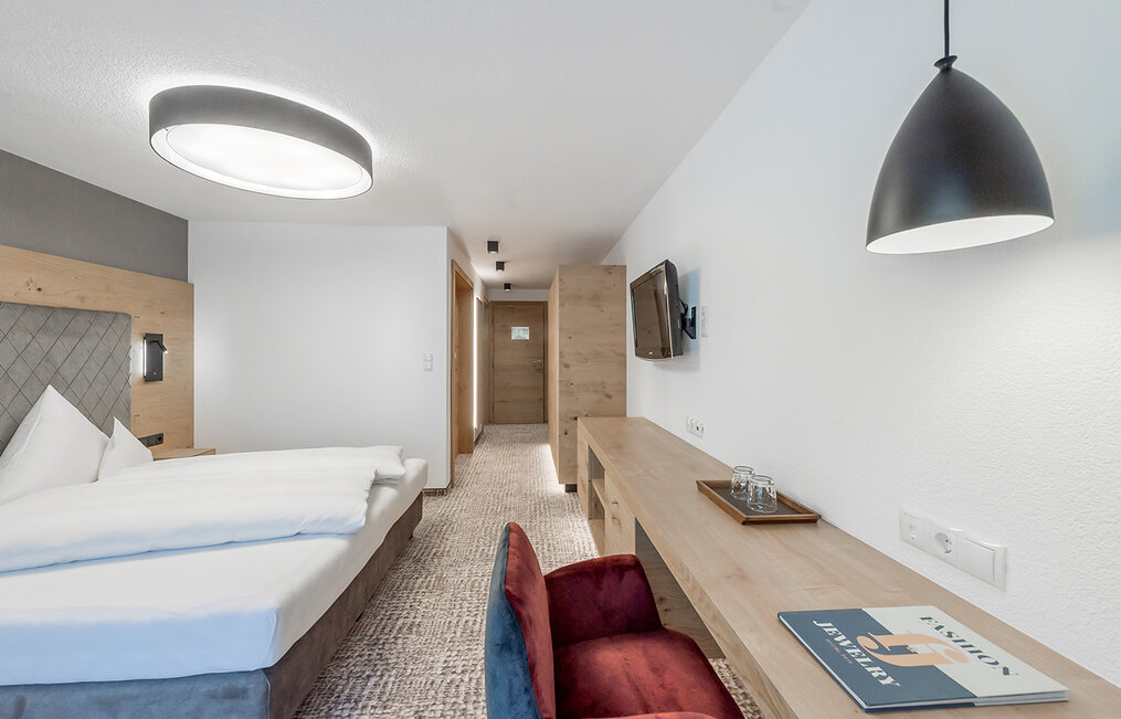Modernes Hotelzimmer mit einem Doppelbett, einem an der Wand montierten Fernseher, einem Schreibtisch aus Holz mit einem roten Stuhl und einer Hängeleuchte. Das Zimmer ist in neutralen Farbtönen gehalten und im minimalistischen Stil gestaltet.