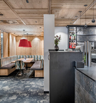 Modern restaurant interior featuring wood panelling, cozy seating booths, patterned carpeting, a red pendant light above a seating area and a bar with bottles and glasses displayed on shelves behind the counter.