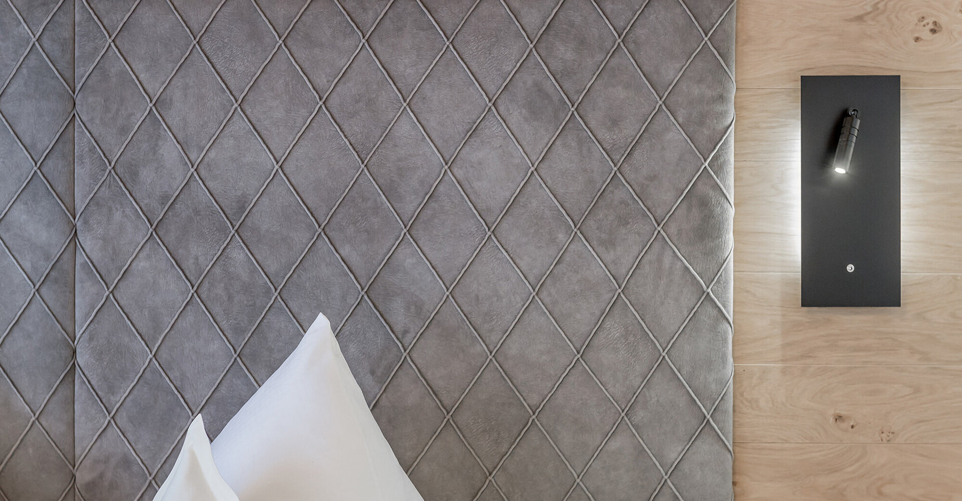 Close-up of a grey, diamond-tufted headboard and white pillows against a light wood wall. A black wall-mounted reading lamp next to the bed is switched on.