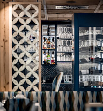 A stylish restaurant interior featuring patterned décor, a glass shelving unit and a shelf behind the bar stocked with various bottles. Tables with white tablecloths are in the foreground.