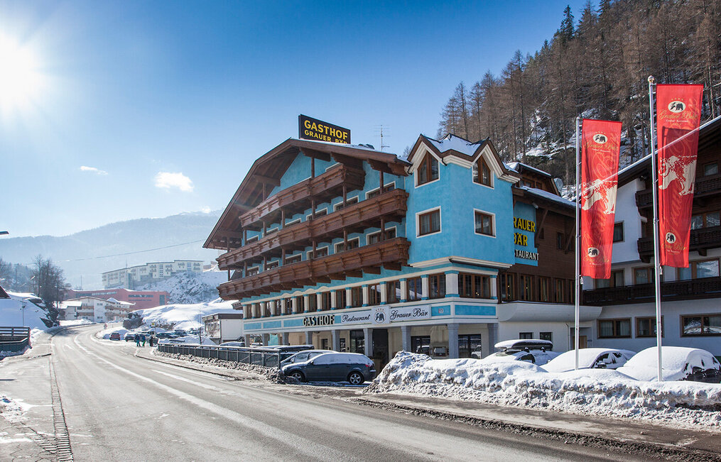 A blue hotel with wooden balconies stands on a snow-covered street beneath a clear sky. Red flags flutter in front of the building, cars are parked along the street. Snow covers the ground and the roofs with mountains in the background.