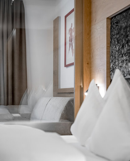 Ein modernes Hotelzimmer mit einem ordentlich gemachten Bett, weißen Kissen, einem grauen Sofa, braunen Vorhängen und einem gerahmten Wandbild. Die Atmosphäre ist hell und minimalistisch.