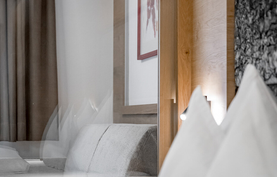A modern hotel room with a neatly made bed, white pillows, a gray sofa, brown curtains and a framed wall picture. The atmosphere is bright and minimalist.