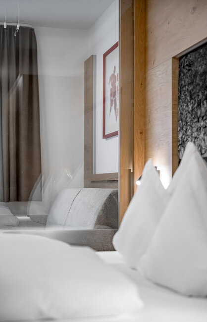 Ein modernes Hotelzimmer mit einem ordentlich gemachten Bett, weißen Kissen, einem grauen Sofa, braunen Vorhängen und einem gerahmten Wandbild. Die Atmosphäre ist hell und minimalistisch.