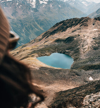 A person looks out at a mountain landscape featuring a heart-shaped blue lake, surrounded by rocky terrain and distant snow-capped peaks.