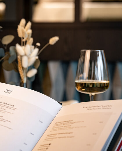 A close-up of an open menu on a table featuring two glasses of white wine and dried flowers in a vase, set against a softly blurred background.