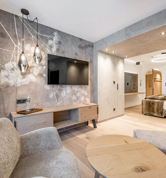Modern hotel suite featuring two gray chairs, a round wooden table, a large TV on a wooden shelf, abstract wall art and pendant lights - with a view into a cozy bedroom in neutral tones and soft lighting.