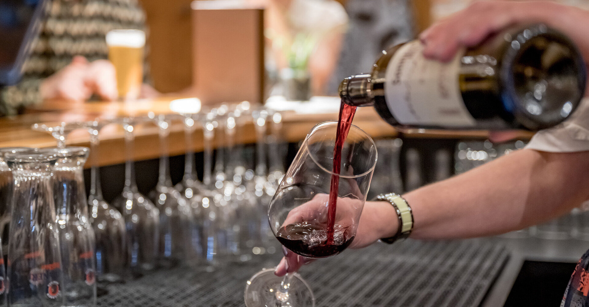 In a bar, someone is pouring red wine into a glass while empty glasses are lined up and people are sitting in the background.