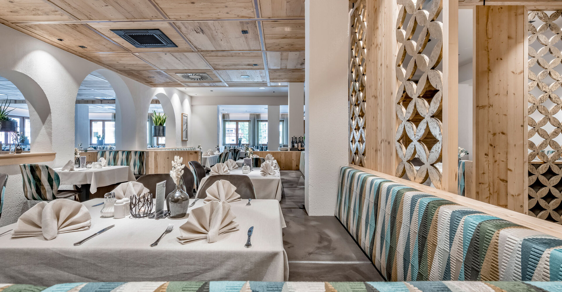A modern restaurant area with white tablecloths, folded napkins, patterned upholstered chairs, wooden elements and decorative partitions, creating a bright and inviting atmosphere.