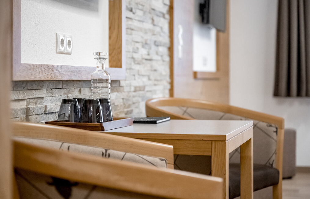 A small wooden table with two upholstered chairs and a tray holding a glass water carafe and glasses, set against a stone accent wall in a modern, brightly lit room.