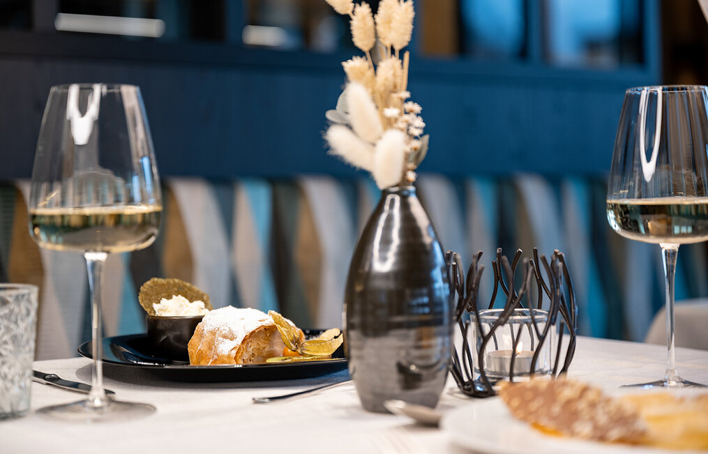 A close-up of a restaurant table featuring two glasses of white wine, a black plate with dessert and whipped cream, a modern vase with dried flowers and elegant tableware.
