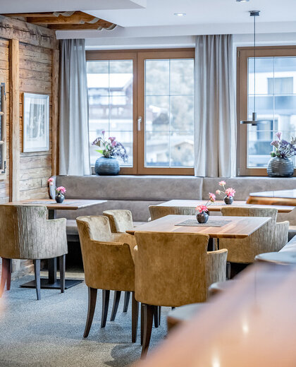 Modern bar area with light brown upholstered chairs, wooden tables and gray sofas. Large windows with gray curtains let in daylight. Vases of flowers adorn the tables and windowsills.