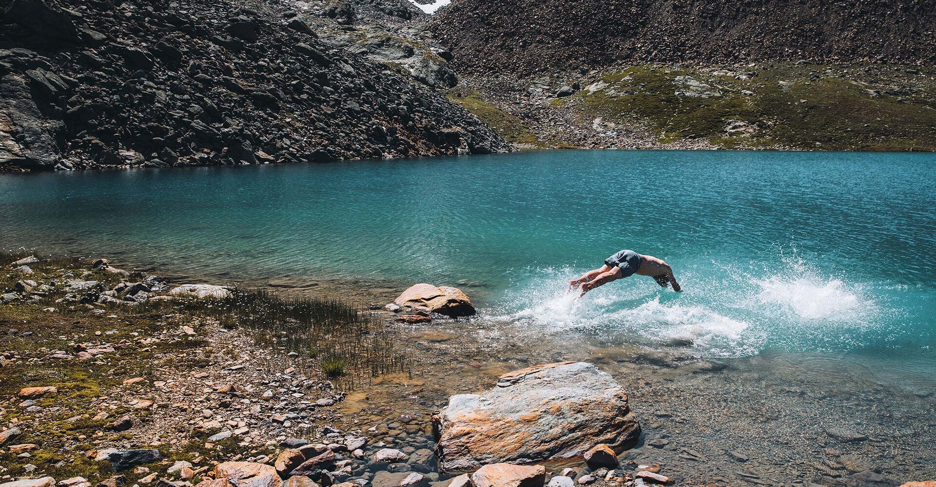 A person in swimming trunks enthusiastically jumps into a clear, blue mountain lake surrounded by rocky terrain and steep slopes.