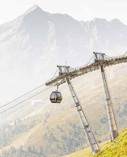 A single gondola cabin travels along a steel rope supported by tall pylons on a grassy mountainside. High mountain peaks rise up in the background.