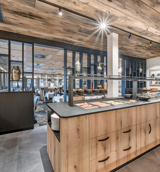 A modern restaurant buffet area featuring wooden cabinets, various food platters behind a protective screen and plates stacked on the counter. Stylish seating and elegant décor are in the background.