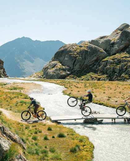 Four cyclists are crossing a small wooden bridge over a mountan brook in a rocky mountain landscape - with one of them lifting the front wheel. A clear sky and distant mountains are in the background.