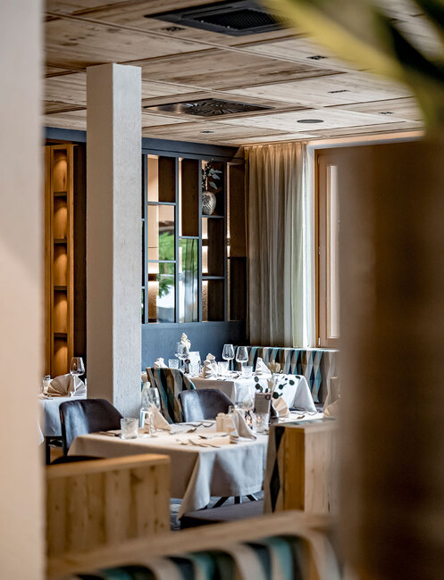 Elegant restaurant interior with neatly set tables, white tablecloths and wine glasses. Warm lighting and wood accents create a cozy, inviting atmosphere. Large windows let in natural light.
