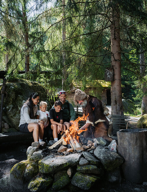 Eine Familie sitzt auf einer Bank neben einem Lagerfeuer im Wald und beobachtet einen Mann in Pelzkleidung, der die Flammen schürt. Hohe Bäume und Felsen umgeben sie und schaffen eine friedliche Atmosphäre in der freien Natur.